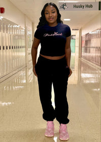 Thumbnail for young woman wearing a black crop top & jogger pants set with 'Pink Seal' branding puff print in school hall with husky sign and lockers in background.