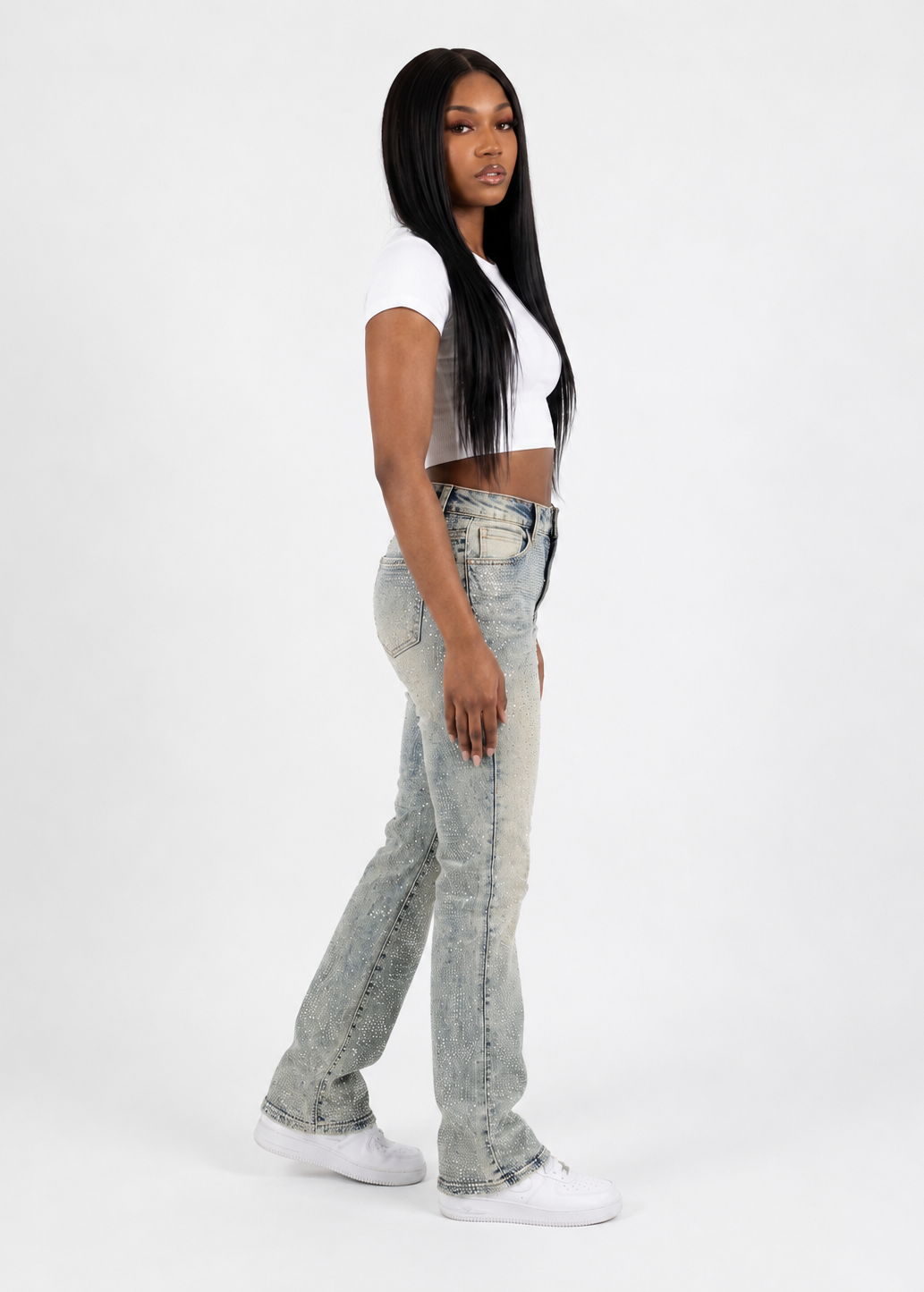 A woman modeling light-wash, high-rise flared jeans with a faded, washed texture, paired with a white crop top and white sneakers, standing against a plain white studio background.
