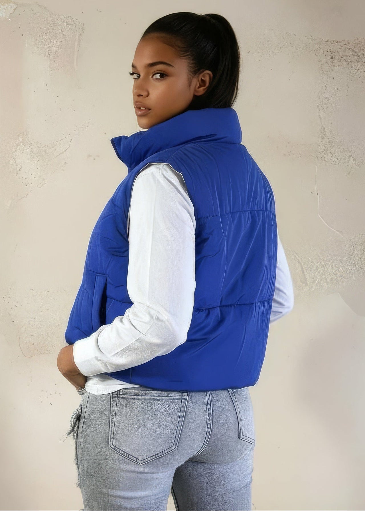 Person wearing a blue puffer vest over a white shirt and gray pants against a beige wall.