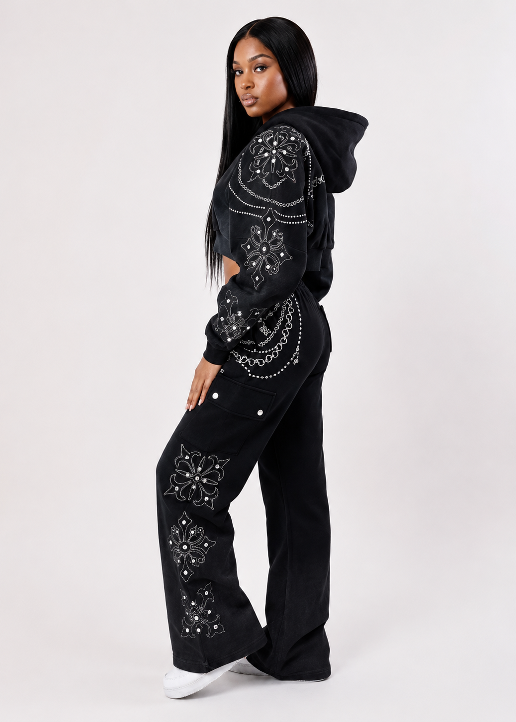 A model wearing a black cropped hoodie and matching wide-leg pants, both decorated with intricate silver rhinestone mandala patterns, posed against a light gray studio background.