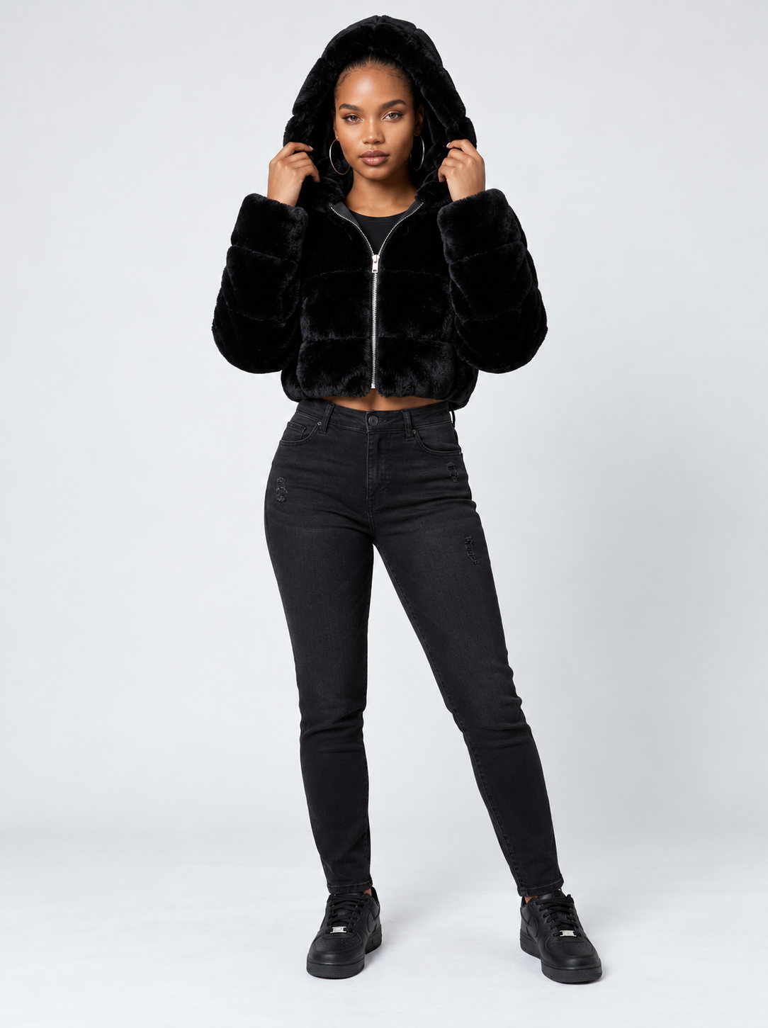 A model wearing a black cropped faux-fur zip-up hoodie with a hood, styled with black skinny jeans and black sneakers, against a light gray studio background.