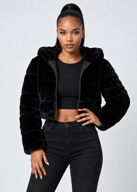 Thumbnail for A woman modeling a black cropped faux-fur jacket with a hood, worn over a black crop top and black jeans, against a light gray studio background.