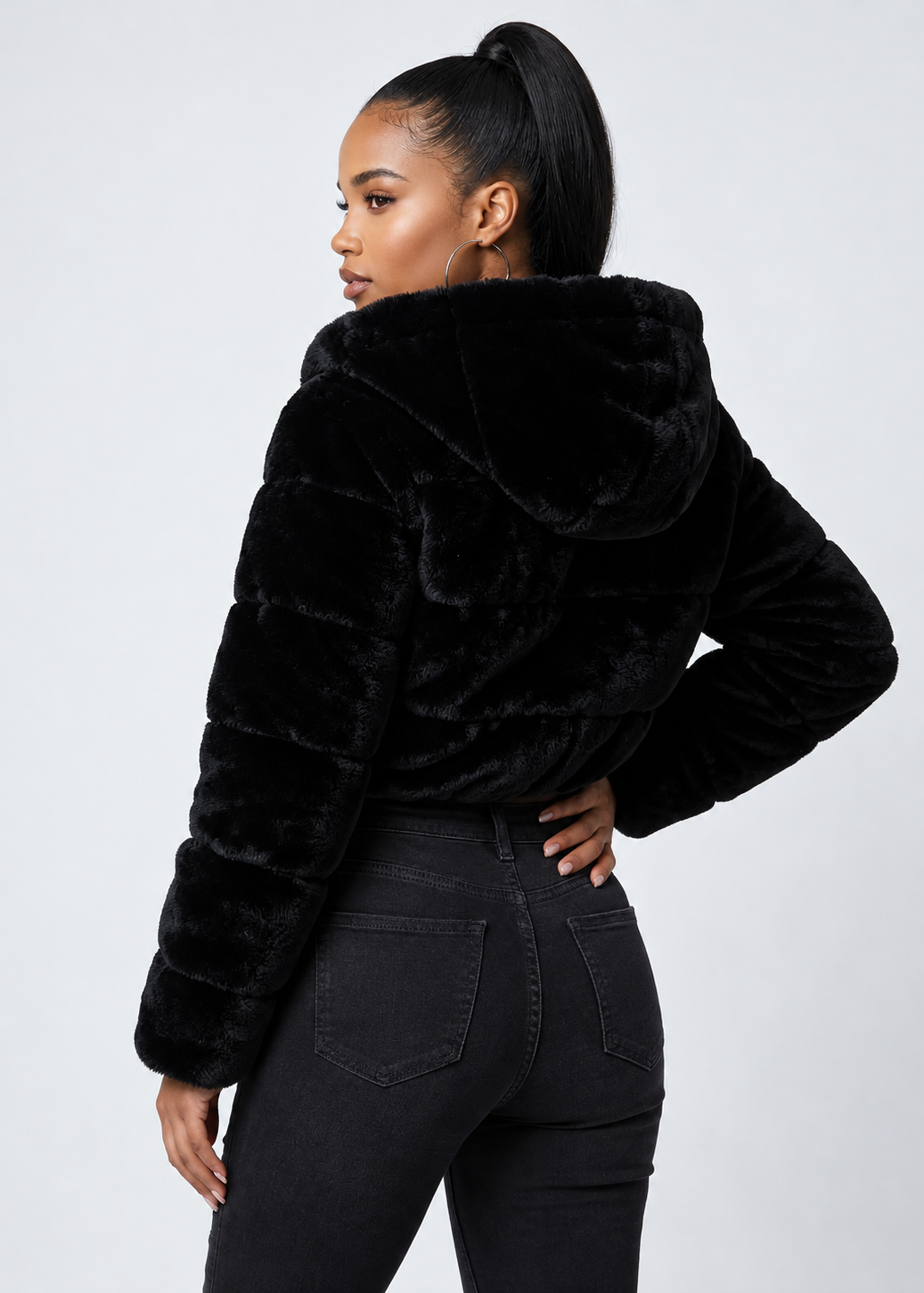A woman shown from the back wearing a black plush faux-fur cropped jacket with a large hood, paired with black jeans, against a light gray studio background.