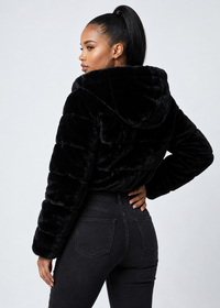 Thumbnail for A woman shown from the back wearing a black plush faux-fur cropped jacket with a large hood, paired with black jeans, against a light gray studio background.