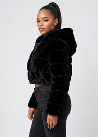 Thumbnail for A woman modeling a black cropped faux fur jacket with a plush hood, paired with black jeans, against a light gray studio background.