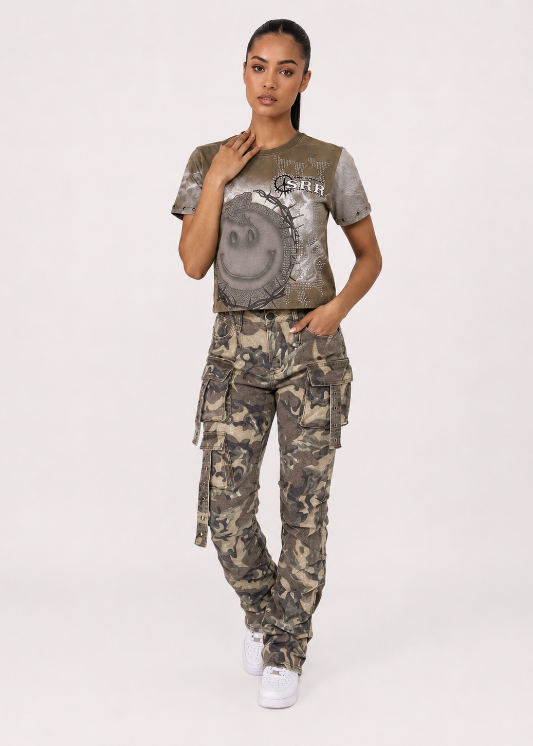 A model wearing a cropped graphic t-shirt and matching camouflage cargo pants, standing against a plain light gray studio background.