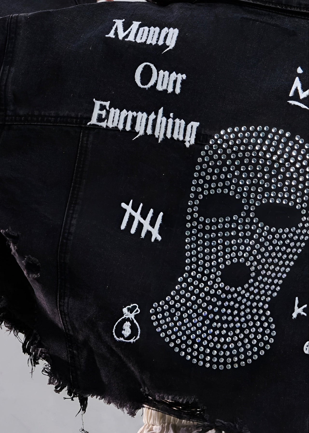 Black denim jacket with 'Money Over Everything' text and rhinestone design on a gray background