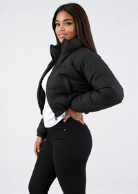 Thumbnail for A woman modeling a black cropped puffer jacket with a high collar, worn over a white top and black pants, against a plain light gray studio background.