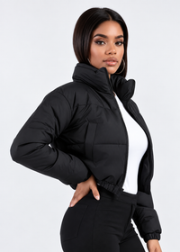Thumbnail for A woman modeling a black cropped puffer jacket with a high stand-up collar and front zipper, worn over a white top and black pants against a light gray studio background.