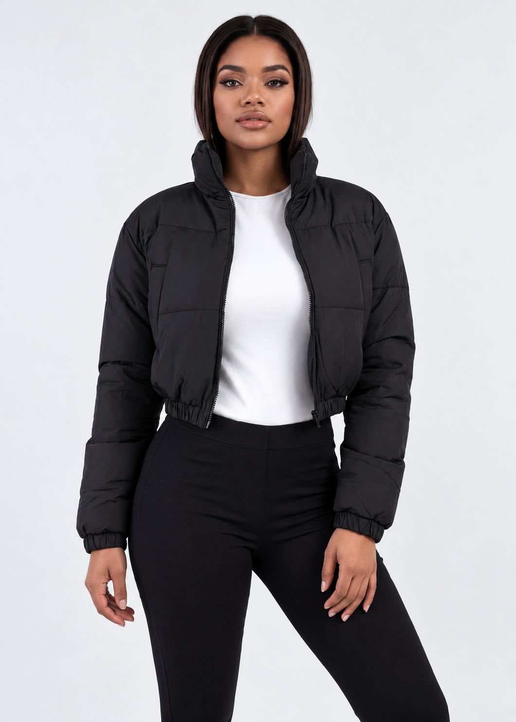 A woman modeling a black cropped puffer jacket over a white top and black pants, photographed against a clean light gray studio background.