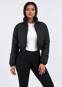 Thumbnail for A woman modeling a black cropped puffer jacket over a white top and black pants, photographed against a clean light gray studio background.