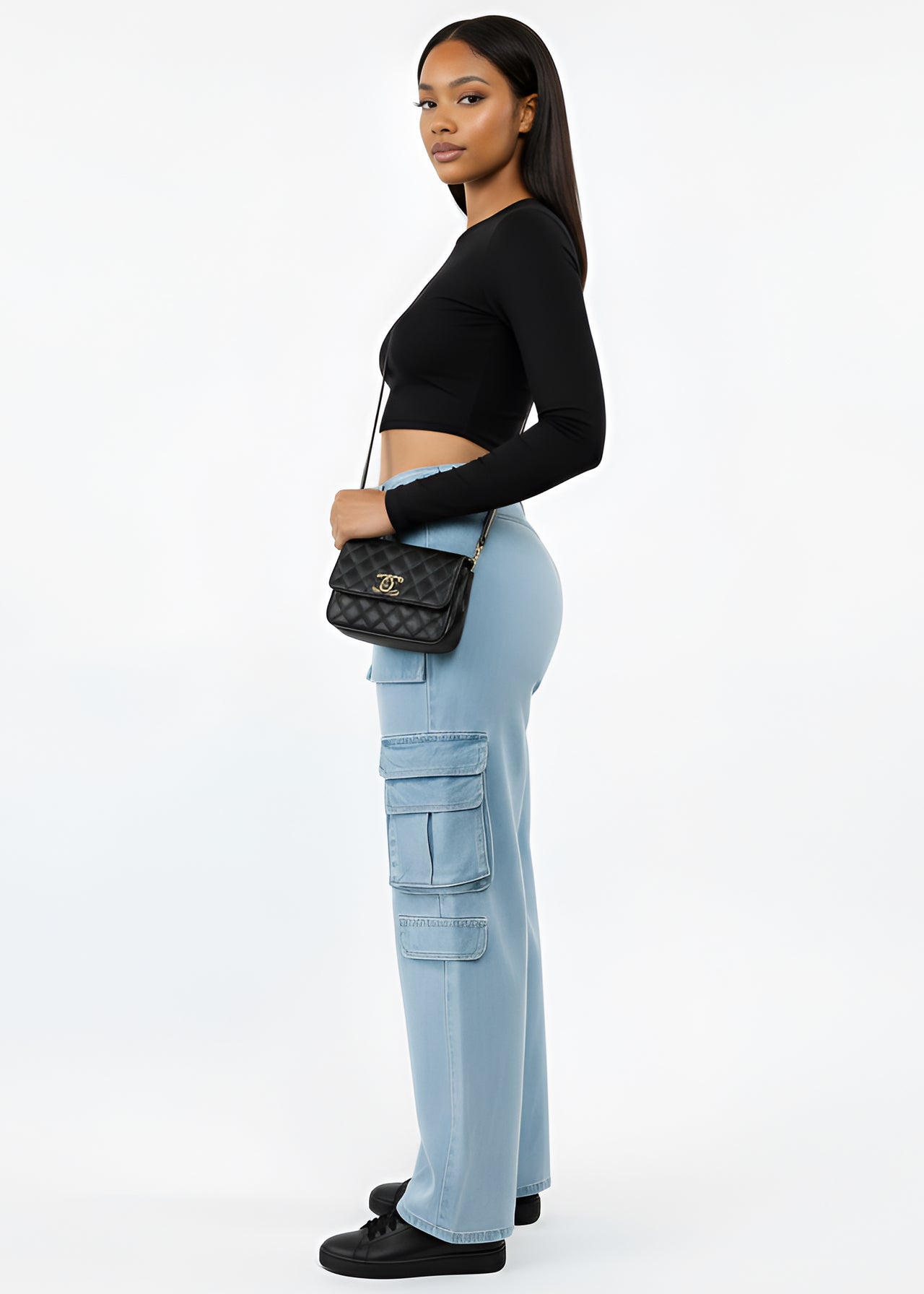 Woman wearing a black long-sleeve top and Pink Seal light blue cargo pants, holding a black handbag on a white background.