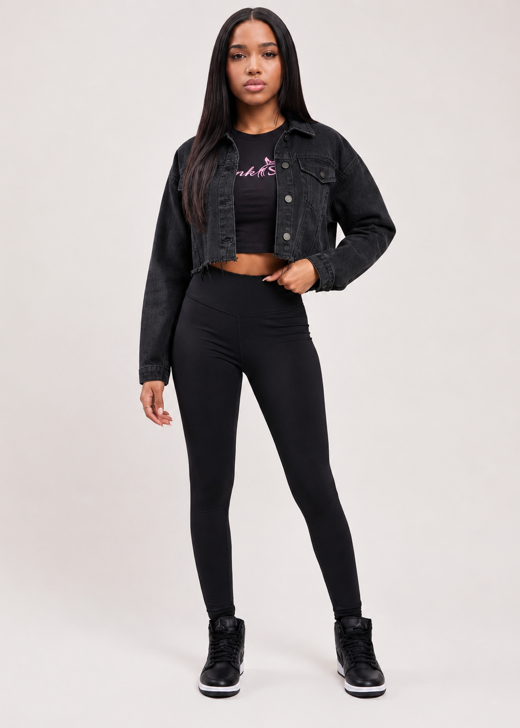 A woman modeling a black denim jacket, black cropped tank top with a pink graphic, black leggings, and black sneakers against a light gray studio background.