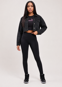 Thumbnail for A woman modeling a black denim jacket, black cropped tank top with a pink graphic, black leggings, and black sneakers against a light gray studio background.