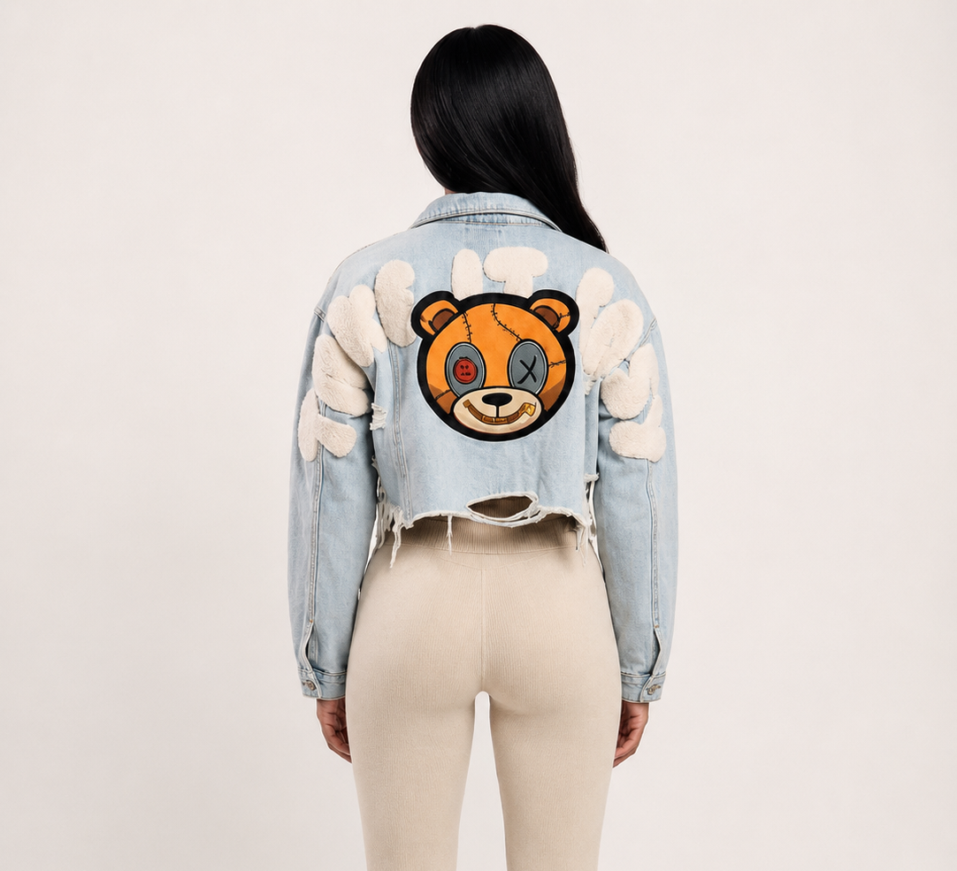 A model shown from the back wearing a light blue cropped denim jacket with a large embroidered teddy bear face patch on the back 