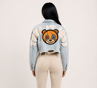 Thumbnail for A model shown from the back wearing a light blue cropped denim jacket with a large embroidered teddy bear face patch on the back 