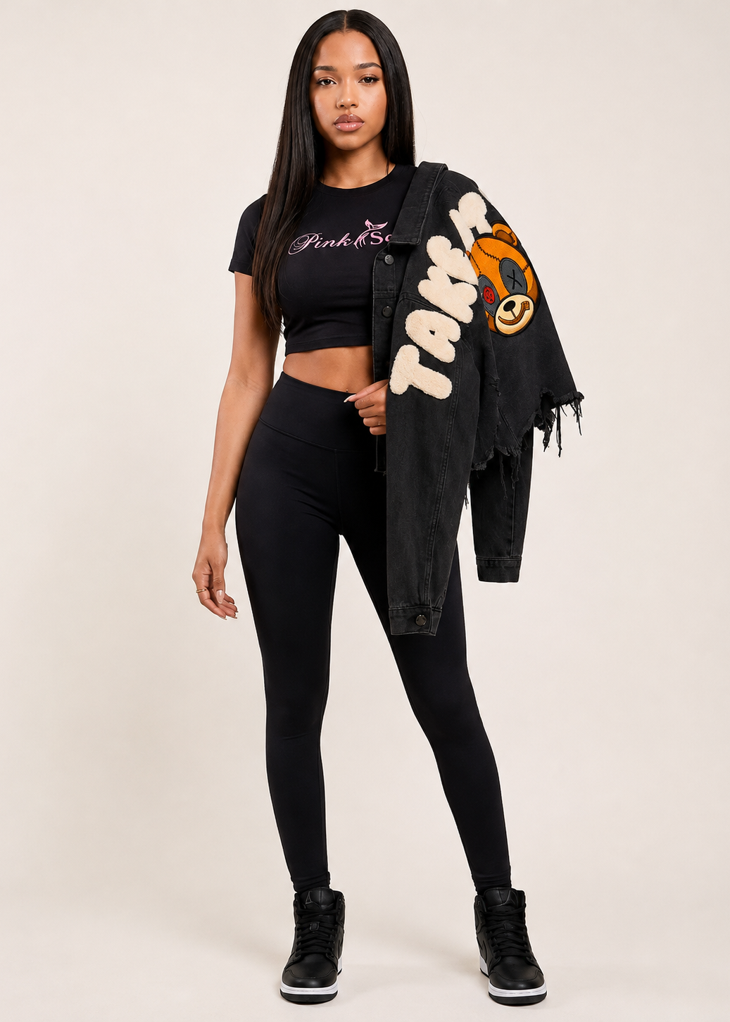 A woman modeling a black short-sleeve crop top with 'Pink Seal' text, black leggings, and a black frayed denim jacket draped over her shoulder 
