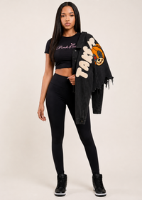Thumbnail for A woman modeling a black short-sleeve crop top with 'Pink Seal' text, black leggings, and a black frayed denim jacket draped over her shoulder 