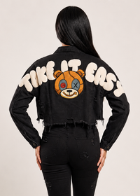 Thumbnail for A model shown from the back wearing a black cropped denim jacket with a distressed frayed hem, featuring a graphic of a teddy bear face and the text 'TAKE IT EASY' in white. 