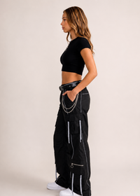 Thumbnail for A woman stands in profile wearing a black short-sleeve crop top, black cargo pants with silver zipper and chain details, a black belt, and white sneakers against a plain light beige studio background.