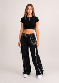 Thumbnail for A model stands against a light beige studio background wearing a black short-sleeve crop top, black wide-leg cargo pants with silver chain and zipper details, and white sneakers.