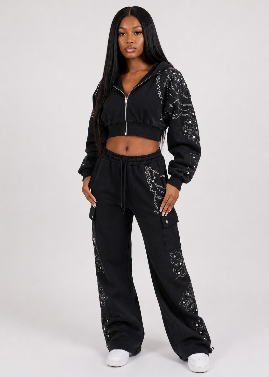 A woman modeling a black two-piece tracksuit with silver chain print details on the sleeves and legs, featuring a cropped zip-up hoodie and matching wide-leg cargo pants, against a light gray studio background.
