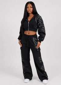 Thumbnail for A woman modeling a black two-piece tracksuit with silver chain print details on the sleeves and legs, featuring a cropped zip-up hoodie and matching wide-leg cargo pants, against a light gray studio background.