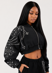Thumbnail for A woman modeling a black cropped zip-up jacket with white embroidered chain and floral patterns on the sleeves, paired with black sweatpants, against a light gray studio background.
