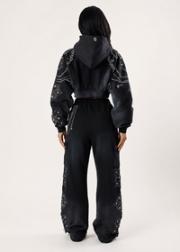 Thumbnail for A model viewed from the back wearing a black cropped hoodie with silver chain embroidery on the sleeves and hood, paired with matching black wide-leg pants with chain detailing down the legs, set against a plain light gray studio background.