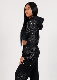 Thumbnail for A woman modeling a black cropped hoodie and matching black cargo pants with intricate silver rhinestone and bead embroidery on the sleeves and upper thighs, set against a plain light gray studio background.