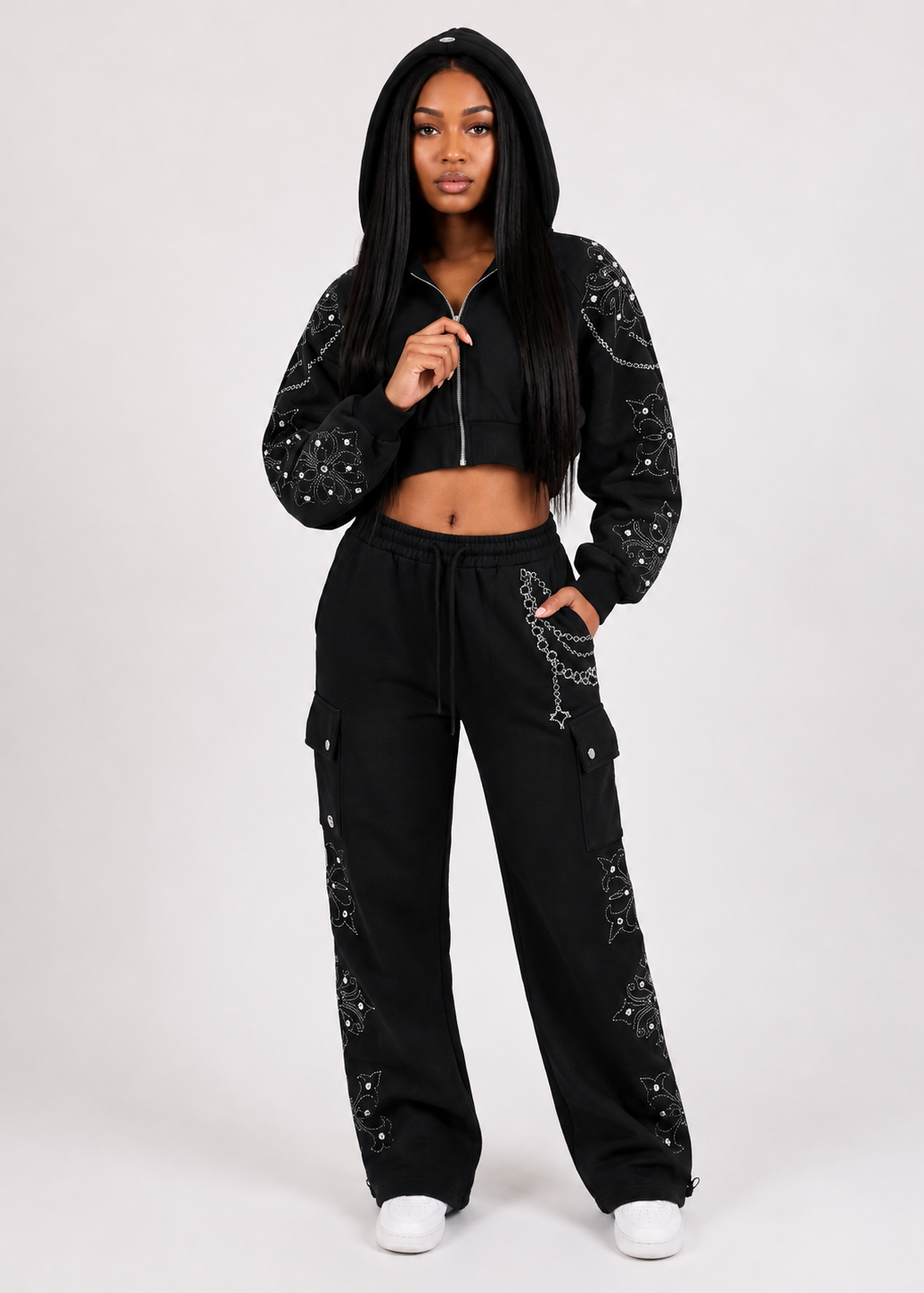 A woman modeling a black two-piece tracksuit with silver rhinestone embellishments on a light gray studio background. The set includes a cropped zip-up hoodie and matching wide-leg cargo pants.