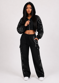 Thumbnail for A woman modeling a black two-piece tracksuit with silver rhinestone embellishments on a light gray studio background. The set includes a cropped zip-up hoodie and matching wide-leg cargo pants.