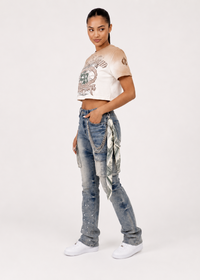 Thumbnail for A model wearing a beige crop top with a graphic print, distressed blue jeans with hanging silver chains and a patterned fabric accent, and white sneakers, photographed against a plain light background.