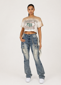 Thumbnail for A model stands against a light gray studio background wearing a beige cropped graphic T-shirt, blue denim cargo jeans with chain details, and white sneakers.