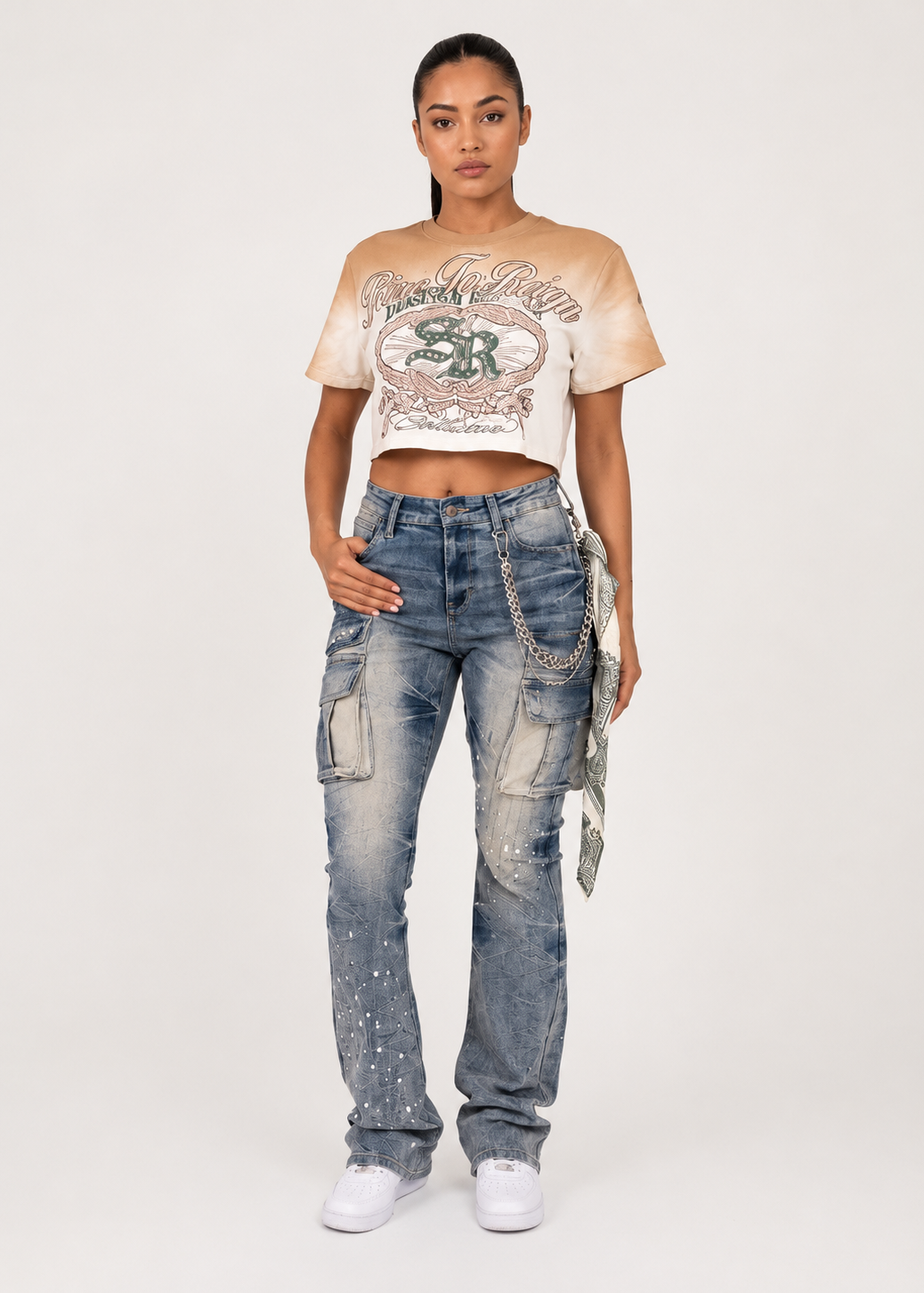 A model stands against a light gray studio background wearing a beige cropped graphic T-shirt, heavily distressed blue jeans with a silver chain belt, and white sneakers.