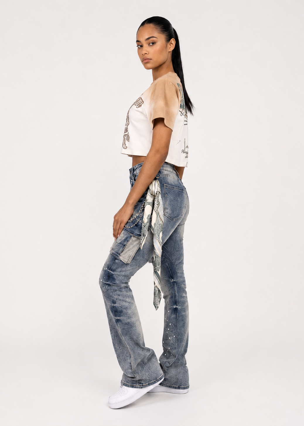 A model is shown from the side wearing a beige cropped t-shirt with a graphic print and high-waisted, wide-leg blue denim jeans featuring a distressed wash and a matching denim tie belt. She is standing against a plain white studio background.